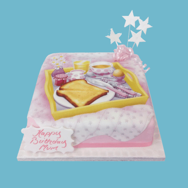 A square cake designed as a whimsical breakfast-in-bed scene, featuring a pink floral bed base topped with edible replicas of toast, a boiled egg, a cup of tea, juice, and a jam jar arranged on a fondant breakfast tray. A personalised message is displayed in a playful font, surrounded by sparkly star toppers and finished with a pink bow. The detailed, realistic design combines charm and elegance, making it a standout centrepiece for birthdays or special occasions. Customisable in flavour and message, the cake offers both visual delight and delicious taste.