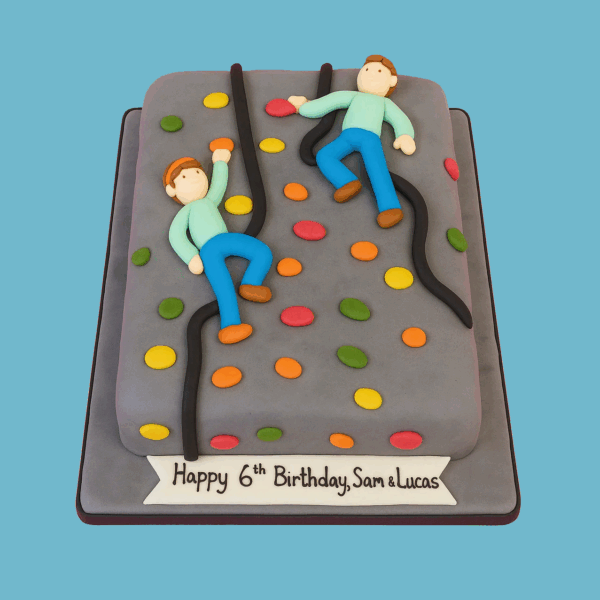 A cake designed to look like a grey climbing wall with a textured surface and brightly colored handholds scattered across it. Two fondant figures in climbing poses cling to the wall using black fondant ropes. A banner at the cake’s base displays a personalized message. The cake captures the excitement of rock climbing and is customizable in size and flavor for celebrations.