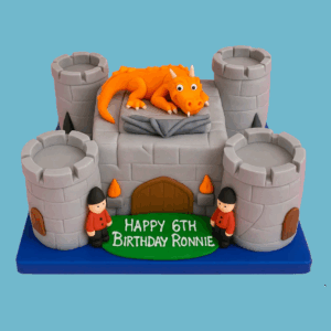 A detailed cake shaped like a grey stone medieval castle with towers and battlements, featuring an orange fondant dragon with spread wings perched on top. Fondant guards stand around the castle, and colorful flame decorations add a fiery effect. The cake sits on a green base resembling grass with a personalized message on the front. Available in various sizes and flavors, this fantasy-themed cake is designed for birthdays and special celebrations.