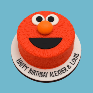 A round cake decorated as Elmo from Sesame Street, with a bright red textured surface resembling fur, large white eyes with black pupils, an orange nose, and a wide black smile. The cake is bordered by a smooth fondant ribbon and includes space for a personalized message, making it a vibrant and cheerful centerpiece for kids’ parties.