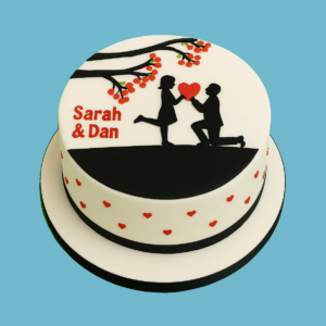 Two-tier Engagement Cake with a smooth fondant finish, decorated in a romantic red and white theme. The top tier features a silhouette of a man kneeling and offering a red heart to a woman beneath a stylised tree branch with red blossoms. The bottom tier is adorned with scattered red hearts, and personalised names appear neatly on the top tier. A ribbon trims each tier’s base. Elegant and symbolic, the cake is designed for engagements, proposals, or anniversaries, with options for custom messages, flavours, and colours.
