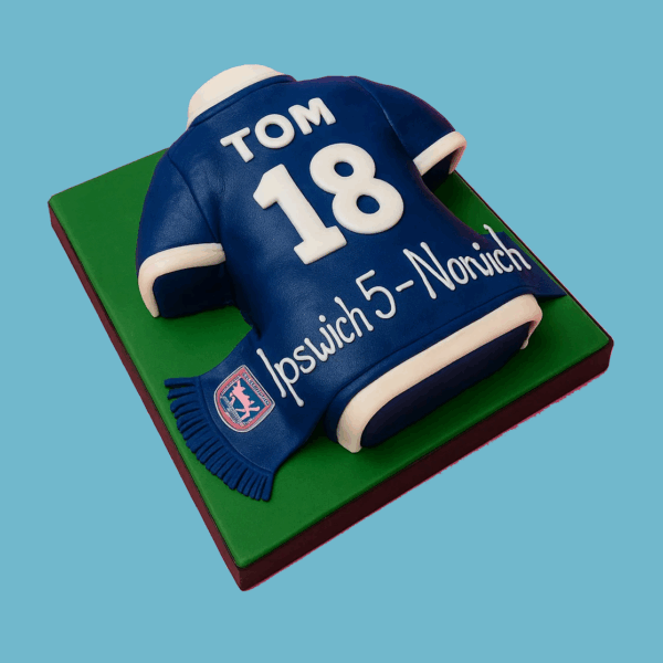 A cake shaped like a classic football shirt with smooth blue and white fondant, featuring crisp color separation and part of a football club’s crest on the sleeve. The shirt cake sits on a green fondant base designed to look like a football pitch, making it a perfect centerpiece for football fans at birthdays or sports events.