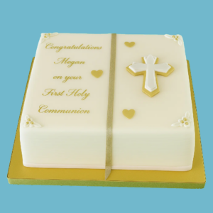 A white fondant cake designed like an open book for a First Holy Communion celebration. The cake features a gold cross in the center, surrounded by gold accents, ribbons, and heart details. Each corner of the book has gold highlights, and there is a personalized plaque on top for a name or message. The overall look is elegant and classic, suitable for a formal yet warm family gathering.