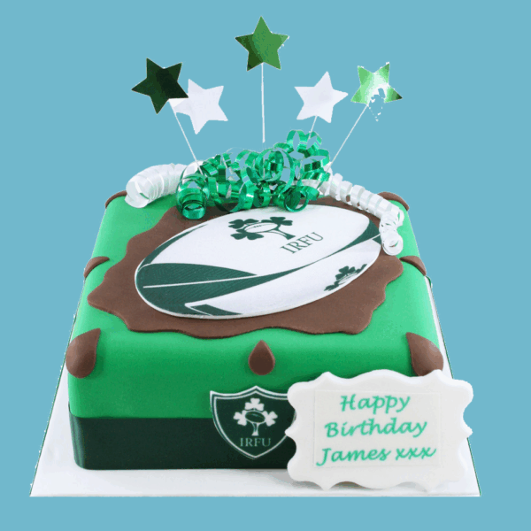 A square cake decorated with vibrant green fondant representing Irish rugby colors, featuring the iconic IRFU logo on top. The cake has a detailed rugby ball design in the center, brown fondant accents on the corners, and a green and white ribbon wrapped around the base. Star-shaped toppers and festive ribbons sit on top, adding a celebratory feel. A customizable edible plaque is included for personalized messages. The cake is designed for rugby fans and special occasions, combining sporty decoration with a festive and joyful appearance.