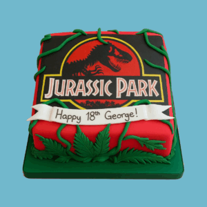 A rectangular Jurassic Park-themed cake featuring the iconic black T-Rex silhouette logo set against a bold red and yellow background. Green fondant vines and leaves frame the edges, creating a jungle-like effect. The cake combines vibrant colours and detailed decorations, making it a standout centrepiece for dinosaur fans and themed celebrations.