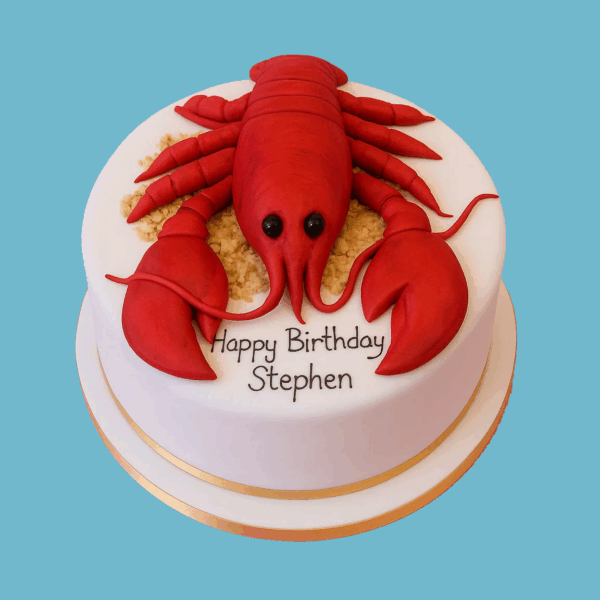 A round Lobster Cake designed to resemble a cooked lobster in vibrant red tones, complete with sculpted claws, legs, and antennae for a realistic appearance. The lobster sits on a smooth white iced base, sometimes decorated with sand-like texture to enhance the seaside theme. This unique and playful cake serves as a standout centrepiece for nautical or seafood-themed celebrations.