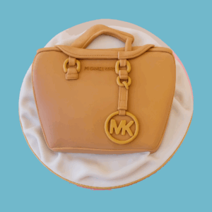 A novelty cake designed to look like a Michael Kors handbag, featuring a smooth light brown fondant exterior with realistic stitching, upright fondant handles, gold-effect buckles, and a hanging "MK" charm. The cake sits on a white fondant base with a draped fabric effect, creating a stylish and elegant centrepiece that mimics a luxury purse with remarkable detail.
