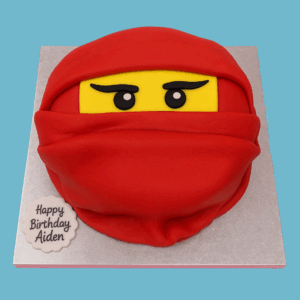 A novelty cake shaped like the head of a red Ninjago ninja character, featuring a bright red mask with bold black and white eyes and a vivid yellow face underneath. The angular design captures the fierce and dynamic look of the Lego Ninjago figure, making it a fun centerpiece for birthday parties and themed celebrations.