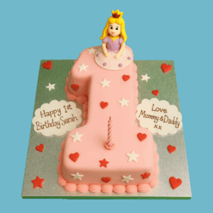 A number one-shaped birthday cake covered in soft pink icing, decorated with red hearts and scattered white stars for a sweet and cheerful appearance. A cute figurine topper sits on the cake, adding a playful touch perfect for a first birthday celebration. The edges are finished with a neatly piped border, and personalised icing plaques display custom messages. This cake serves as a charming centrepiece for milestone events, combining thoughtful design with joyful details.