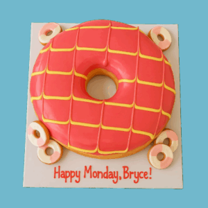 A round novelty cake shaped like a giant pink iced doughnut with a large hole in the center, decorated with white and yellow icing stripes in a playful pattern. Surrounding the main cake are smaller fondant mini doughnuts, adding charm and detail. The cake’s bright colors and whimsical design make it a fun centerpiece for parties and celebrations.