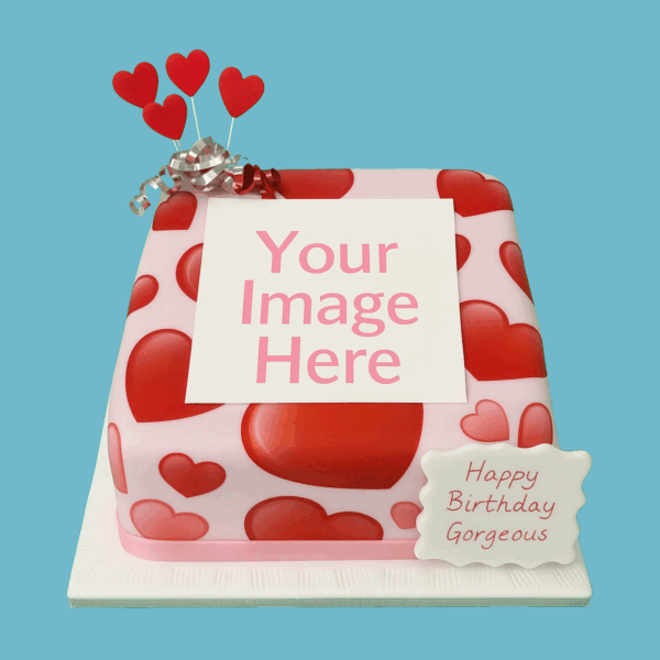 Square pink fondant cake featuring a central photo area framed by red heart decorations. The cake is topped with heart-shaped toppers and wrapped with a silver ribbon at the base. A customisable plaque allows for a personal message. Suitable for romantic or special celebrations like birthdays, anniversaries, or Valentine’s Day.