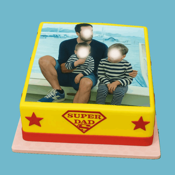 A vibrant, personalised photo cake featuring a “Super Dad” design, decorated with yellow fondant, red accents, and star-shaped toppers. The cake displays a chosen photo beside a bold “Super Dad” graphic, with a red ribbon around the base and bright stars at each corner. A custom message plaque sits on top, allowing space for a personal greeting, making the cake a festive and heartfelt centrepiece for celebrating a father figure.