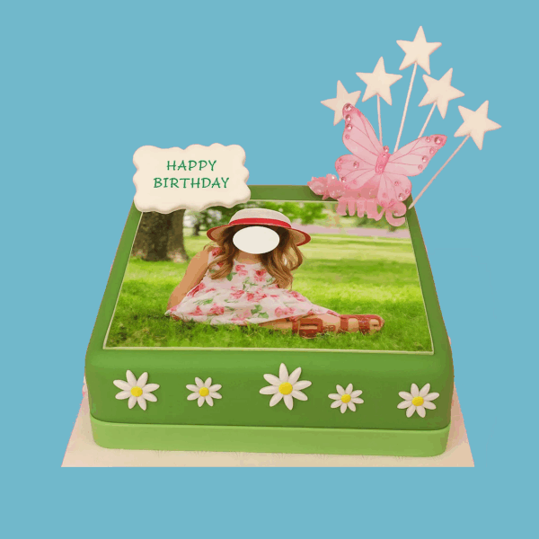 A square cake covered in rich green fondant decorated with delicate white daisy flowers around the base. On top, a pink butterfly rests gently, surrounded by small star-shaped toppers. The center of the cake features a space for a personalized photo, framed by floral decorations. An edible plaque for a custom message is placed on the side. The cake combines elegant floral and butterfly accents with a festive design, perfect for birthdays, anniversaries, or special celebrations.