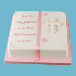 A pink and white First Holy Communion cake designed in the shape of a traditional book with a cross at its center. The cake features a white fondant finish, soft pink ribbons wrapped around the base, and small pink heart decorations. It includes a personalized message space and can be customized with the date of the celebration. The cake is available in various flavors and sizes, making it an elegant and meaningful centerpiece for families celebrating a child’s First Holy Communion.