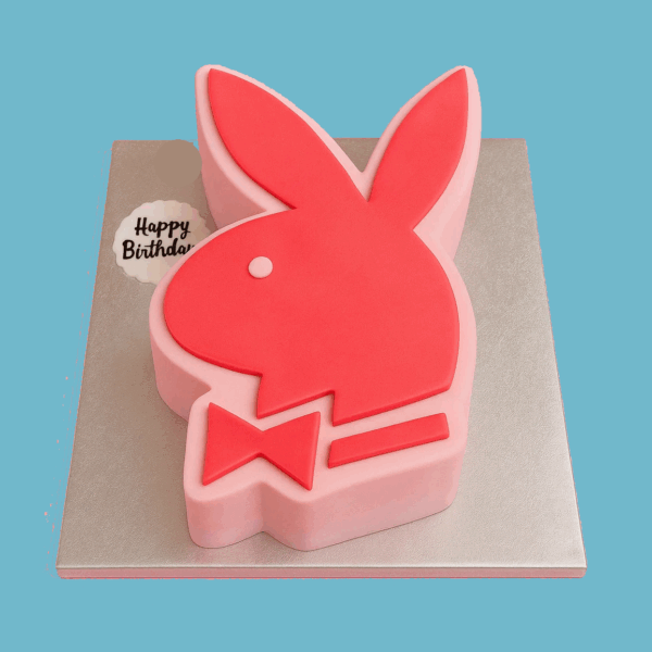 A novelty cake shaped like the iconic Playboy bunny logo, featuring a red silhouette with long upright ears and a sharp bow tie. The bunny is outlined in sleek black for emphasis and sits atop a pink base that contrasts vividly with the red. The design is clean, bold, and instantly recognizable, making it a striking centerpiece for parties or themed events.