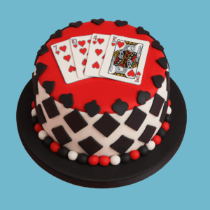A two-tier round cake designed for poker-themed celebrations. The top tier is covered in bold red fondant and decorated with fondant playing cards arranged like a winning hand. The bottom tier features a black and white diamond pattern. Red, white, and black fondant ball borders decorate the edges of both tiers, resembling poker chip colors. The cake presents a polished, elegant look, making it an ideal centerpiece for poker nights or casino-themed parties.
