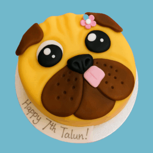 A round novelty cake designed to look like a pug’s face, featuring detailed wrinkles, large dark eyes, a centered black nose, and soft brown floppy ears. A small pink fondant flower decorates the top, adding a cute touch. The cake captures the pug’s charming and lovable expression, making it an appealing centerpiece for dog lovers and themed celebrations.