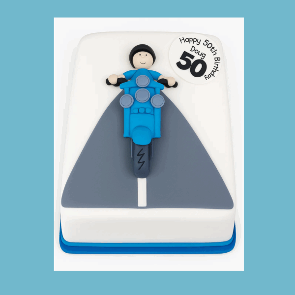 A square cake featuring a stylized blue scooter with silver accents on a grey fondant road atop a white cake surface. A small, detailed figure rides the scooter, adding charm and movement to the scene. The cake is designed as a lively centerpiece suitable for celebrations, customizable in flavor, size, and decoration.