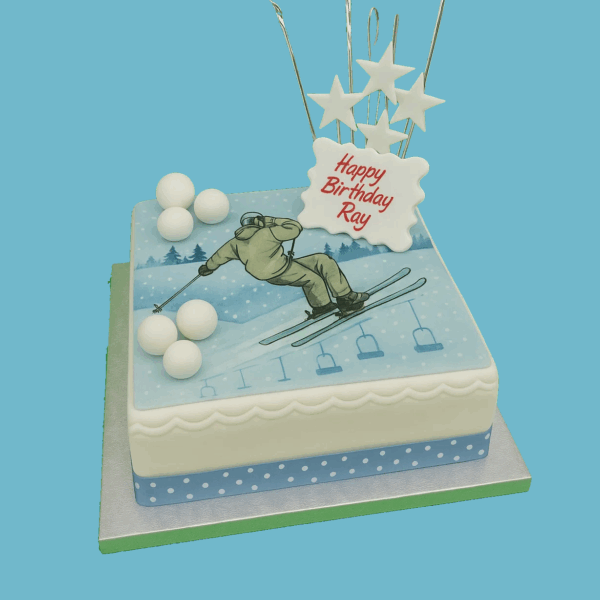 A square cake decorated with blue and white fondant depicting a winter skiing scene. A detailed skier figure is shown skiing down a snowy slope made of white fondant, set against a blue fondant sky. The cake is adorned with small white snowball-like decorations and sparkling star toppers. An edible plaque for a personalized message is placed on the cake. The design captures the excitement of winter sports, making it a festive centerpiece for birthdays, winter parties, or sports events.