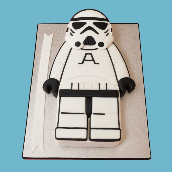 A cake designed as a Star Wars Stormtrooper in the style of a Lego figure, featuring white fondant armour with black detailing on the helmet, chest, and limbs. The blocky shape mimics a Lego character, with square shoulders and firm stance. The cake sits on a silver board that highlights the bold contrast of the design. A playful and detailed creation perfect for Star Wars or Lego-themed celebrations.