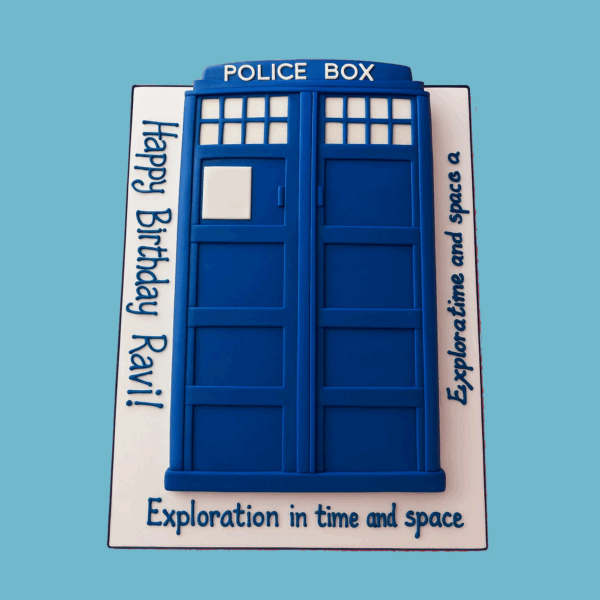 A cake shaped like the iconic blue TARDIS from Doctor Who, featuring sharp edges, white-paned windows, panelled doors, and the “POLICE BOX” sign at the top. The cake has realistic texture and a fondant light on top, standing on a smooth base board. The detailed icing work creates depth, making it a striking centerpiece for Doctor Who fans of all ages.