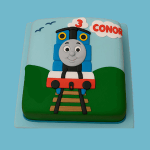 A novelty-shaped Thomas The Tank Engine cake featuring a bright blue engine with Thomas’s smiling cartoon-style face, black wheels, and a red and white carriage attached behind. The cake rests on a brown edible track with a countryside-themed background, including green fields and a light blue sky. Crafted with vibrant colours and smooth detailing, the cake creates a playful and cheerful centrepiece perfect for children’s parties and train-themed celebrations.