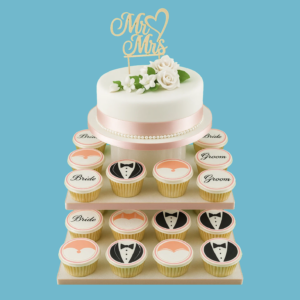 A classic tiered wedding cake with smooth white fondant and delicate floral decorations, topped with a “Mr & Mrs” sign. Surrounding the cake are cupcakes decorated with “Bride” and “Groom” designs, arranged to complement the elegant and charming wedding dessert display.