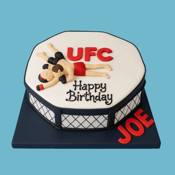 A cake shaped like the UFC octagon fighting arena, with a white octagonal top and black-and-white fondant fence details around the sides. Two fondant figures representing fighters are locked in combat on top. The cake sits on a sleek black rectangular base that can be personalised with names or messages, making it a striking centerpiece for fight fans and sports celebrations.