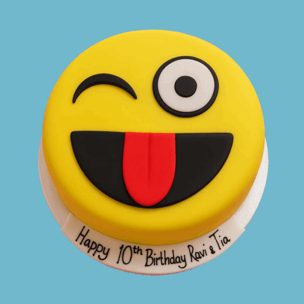 A round cake designed as a winking emoji with a bright yellow surface, featuring one large black eye and one winking eye. Below the eyes is a wide black smile with a red tongue sticking out, giving the cake a playful and mischievous expression. The cake has a space at the base for a personalized message on fondant, making it a colorful and fun centerpiece for celebrations.