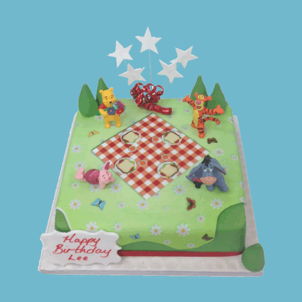 Square Winnie the Pooh celebration cake decorated with a picnic scene featuring Pooh, Piglet, Tigger, and Eeyore on a green meadow with a checkered blanket and colorful flowers. Includes star-shaped toppers and a front plaque for a personalised message.