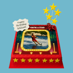 A round celebration cake decorated with bright red fondant and shimmering gold stars. A personal photo is featured at the centre, framed by a stage-like gold star design. Star-shaped toppers stand on top of the cake, and a personalised message plaque sits at the front. The cake has a scalloped edge for a refined finish, creating a bold and festive centrepiece ideal for birthdays or special achievements.