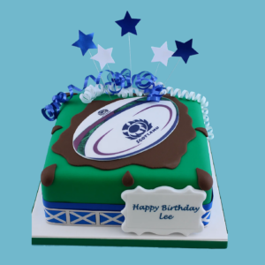 Scotland rugby team cake with a rugby ball featuring the thistle logo on a green pitch-like base, decorated with blue and white stars, ribbons, and a kilt-inspired border.