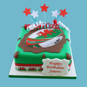 Wales rugby team cake with a rugby ball topper featuring the Welsh dragon and “Cymru” text, decorated in red, white, and green.